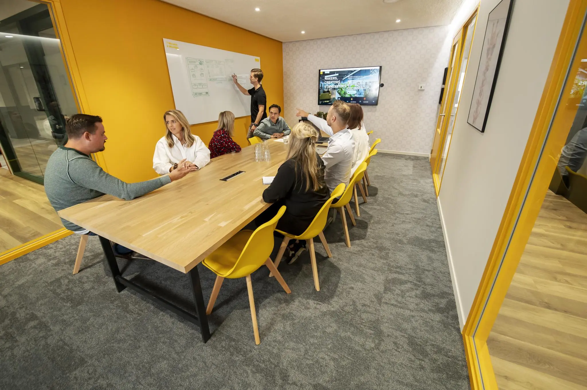 Zeven mensen zitten in een moderne vergaderruimte aan de Aert van Nesstraat terwijl iemand iets uitlegt op een whiteboard.